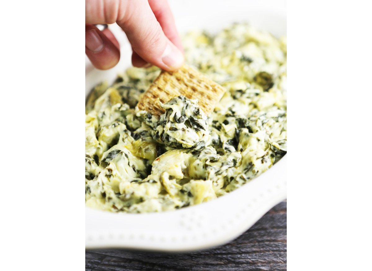 Hand dipping cracker into a dish filled with spinach artichoke dip.