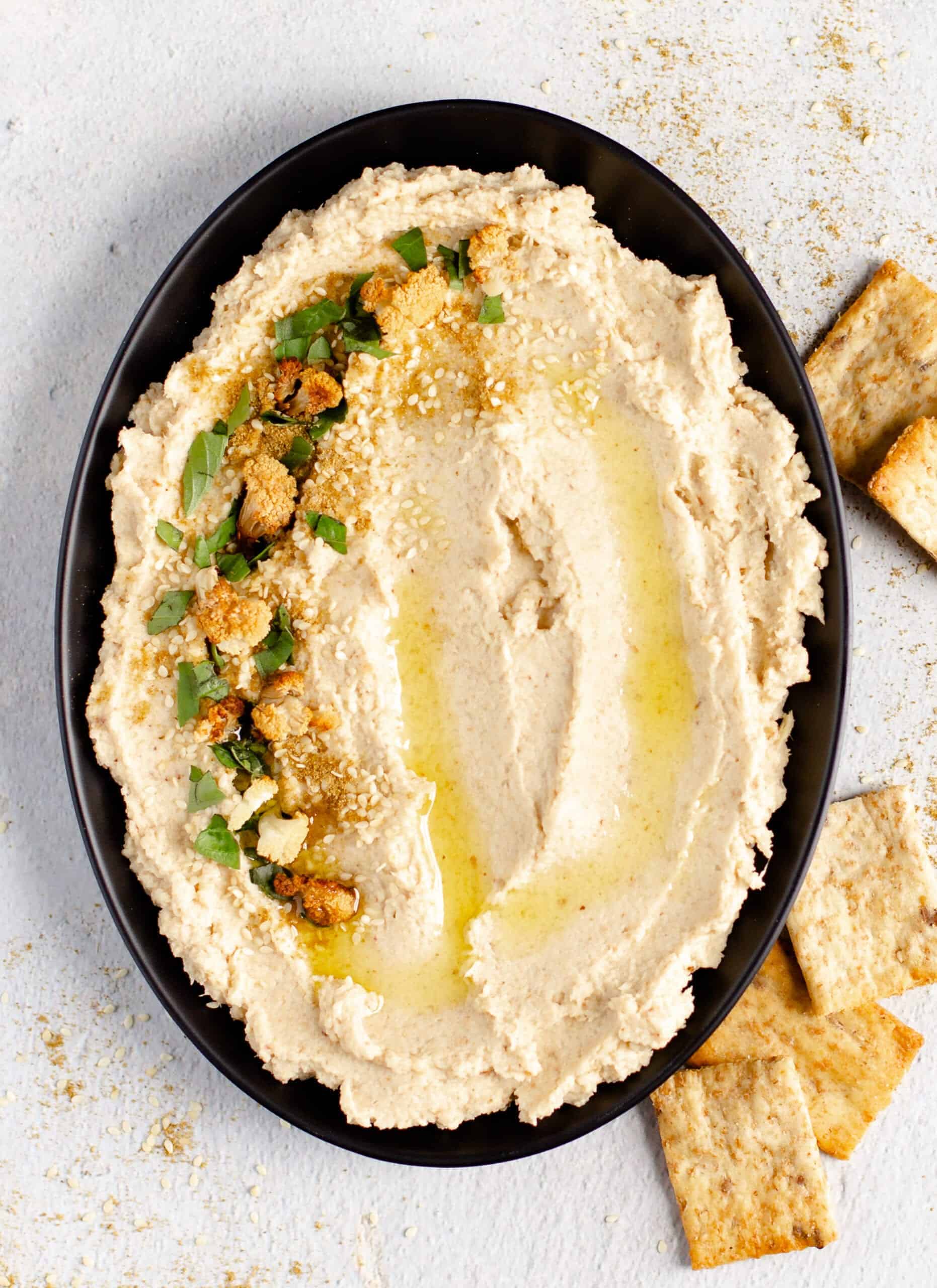 Cauliflower hummus with roasted pieces of cauliflower on a plate with crackers alongside it. 