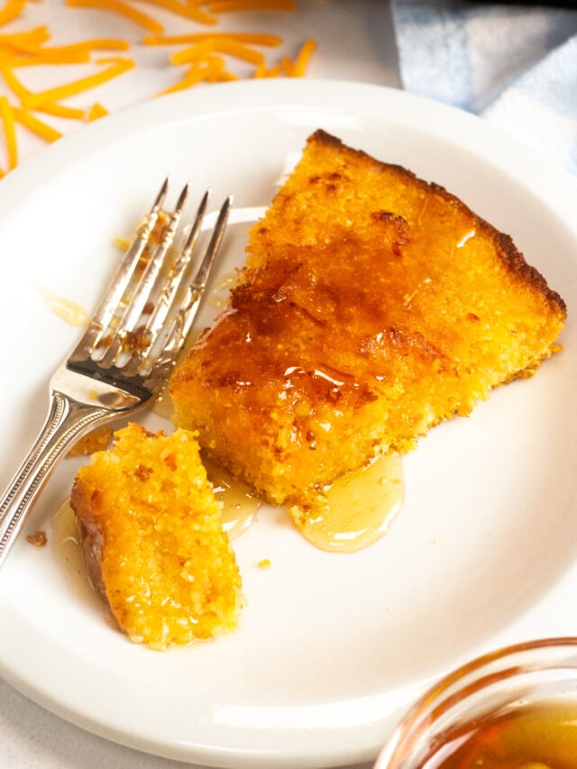Slice of cornbread on a plate with honey drizzled over the top with a fork on the plate. 