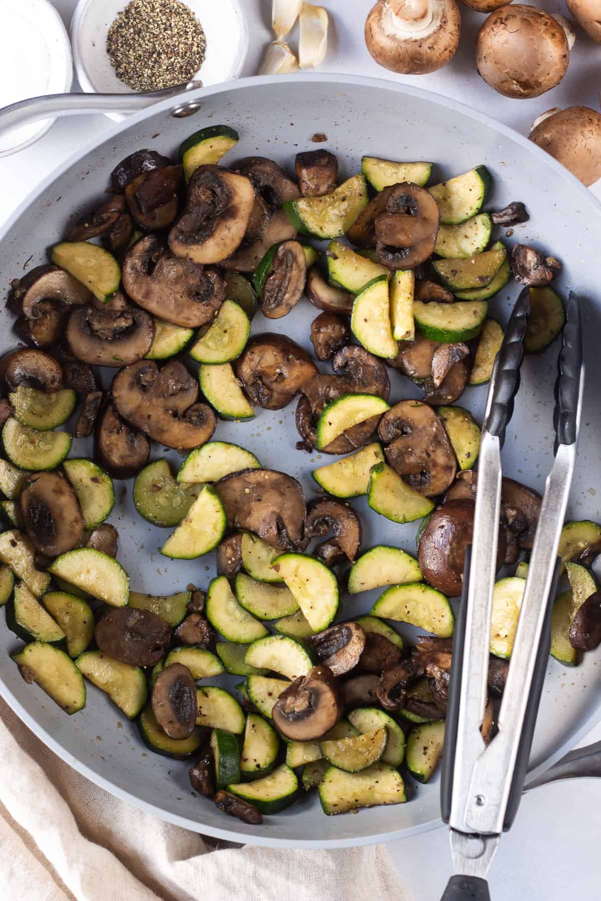Sauteed zucchini and mushrooms in a bowl with a tongs on the side. 
