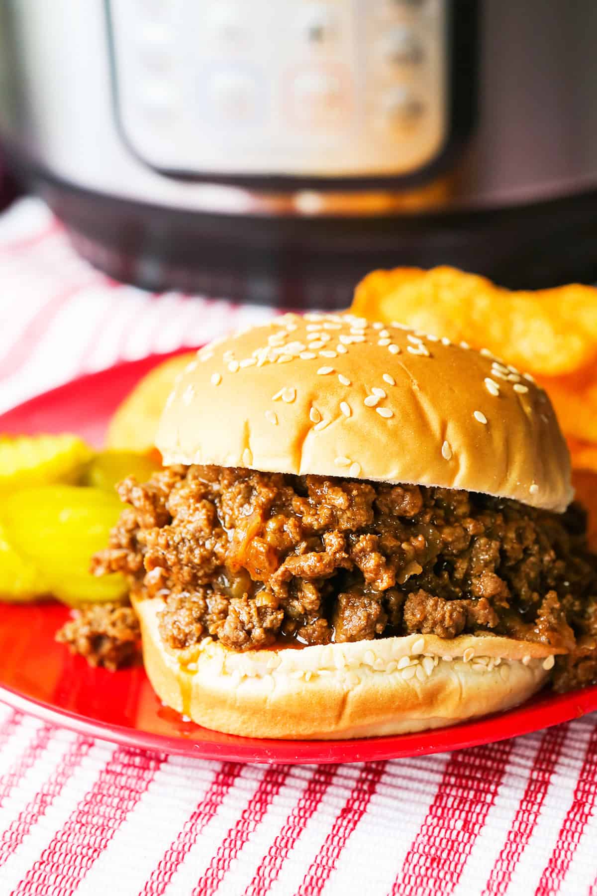 Loaded sloppy joe sandwich on a plate with pickles slices, all of it sitting next to a pressure cooker.