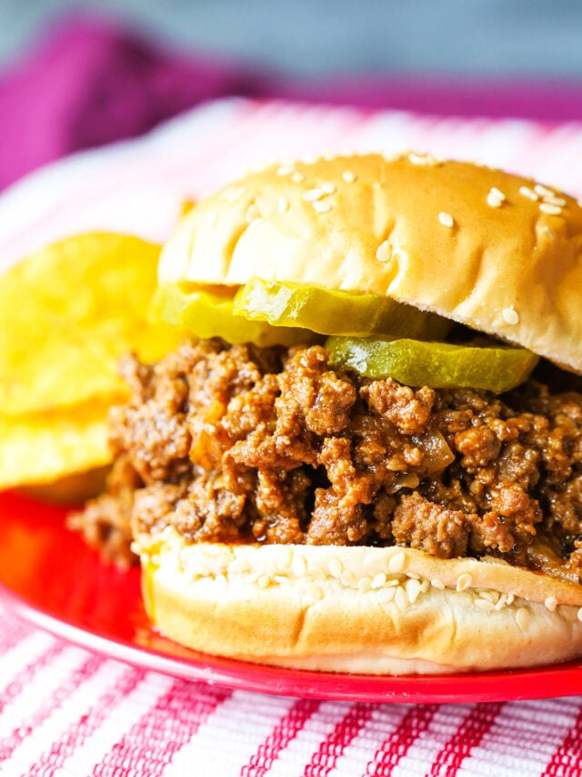 Sloppy joes on a sesame seed bun on a palte with dill pickles on top and chips on the side. 