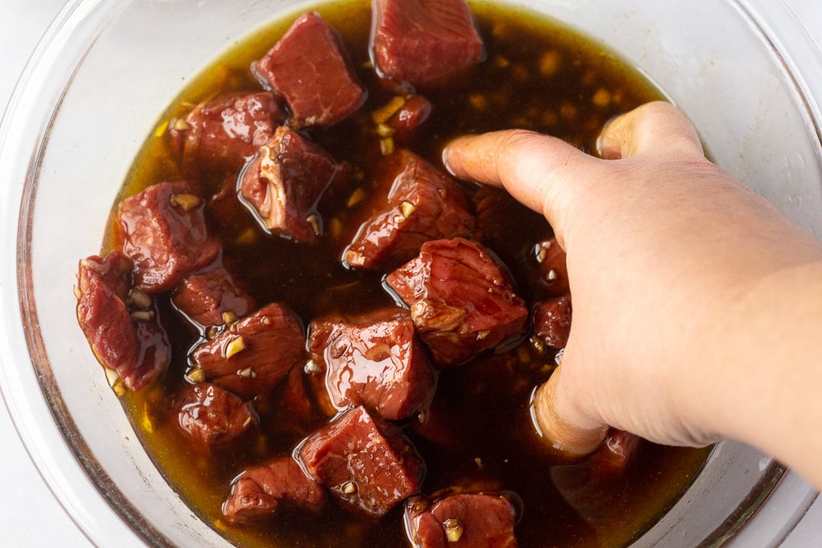 Hand in a bowl tossing around steak pieces in a marinade.