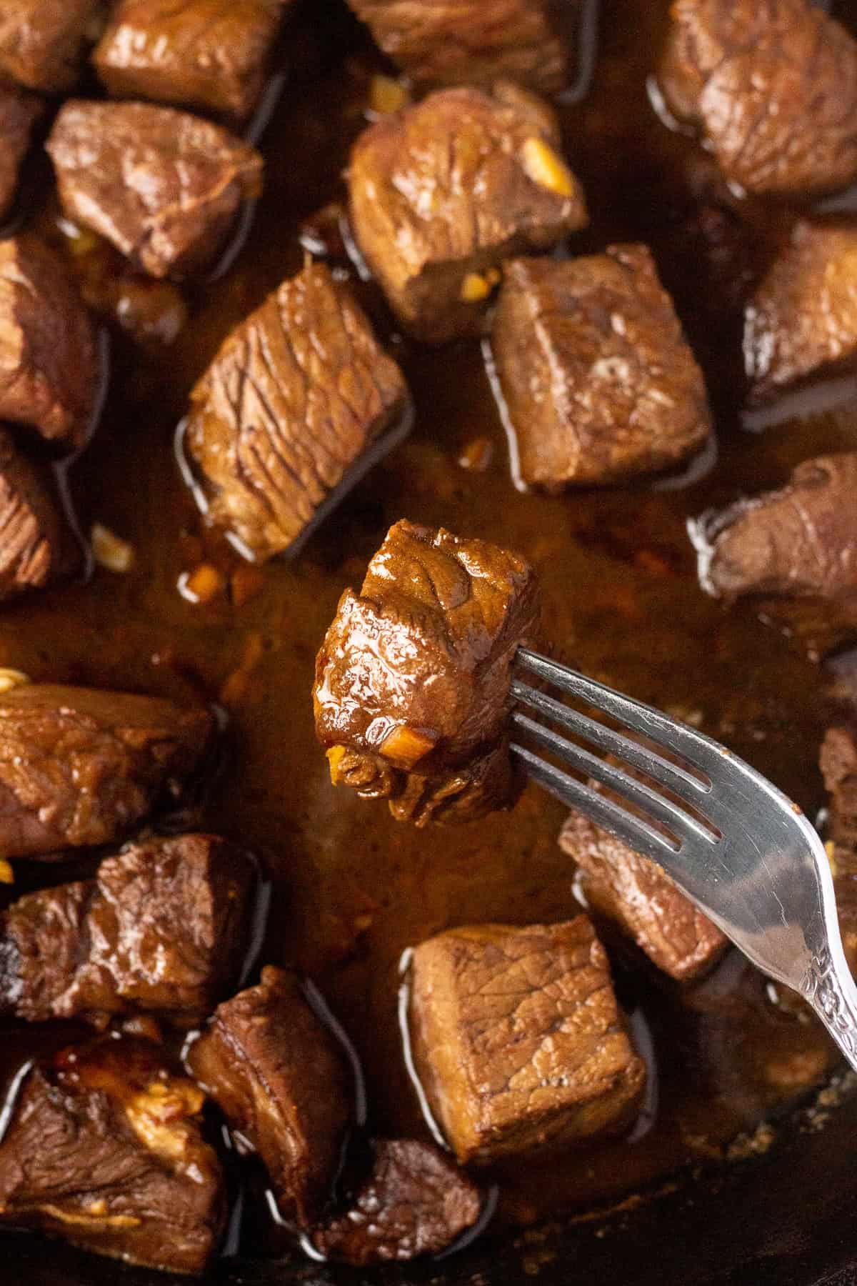 Fork holding up a steak bite, from a pan filled with juicy-looking bites.