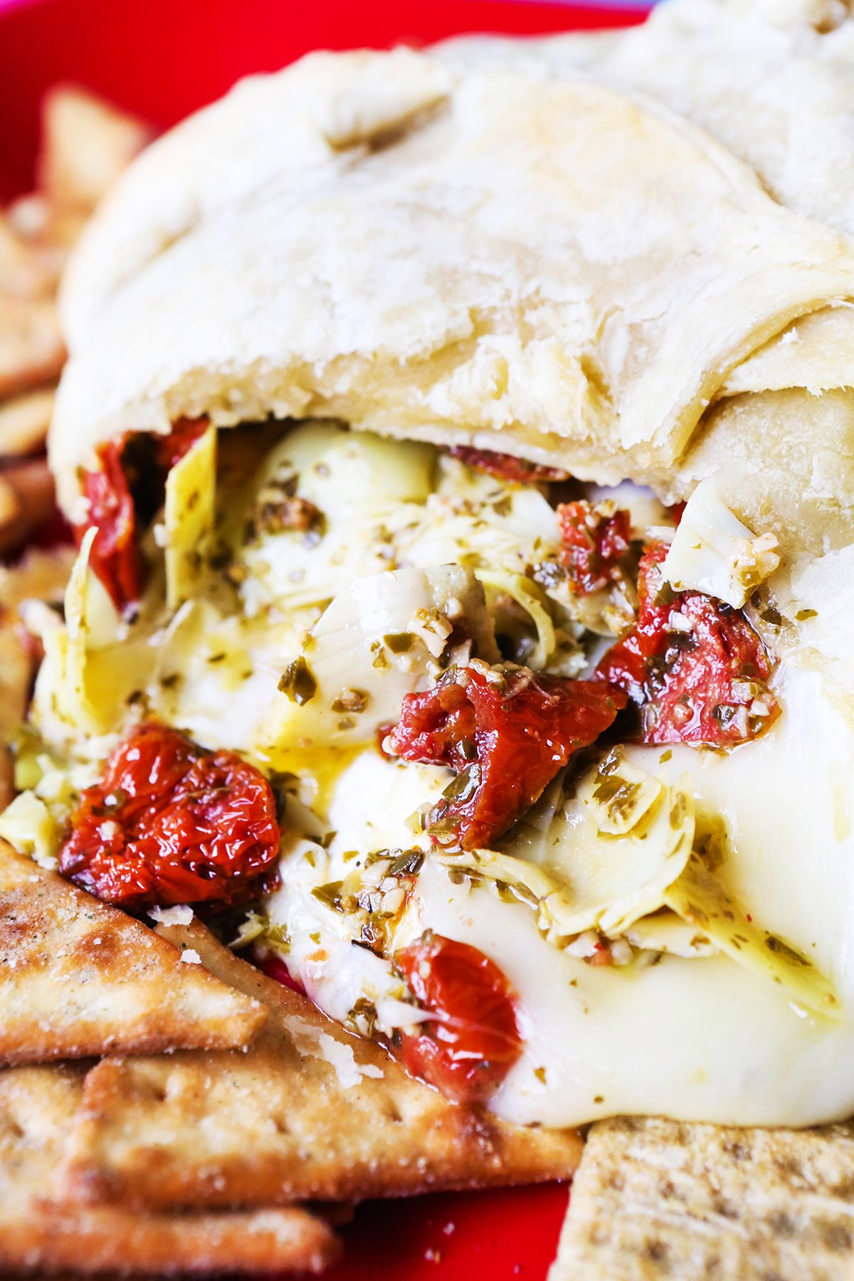 Baked brie with cheese, artichokes and sundried tomatoes spilling out next to crackers. 