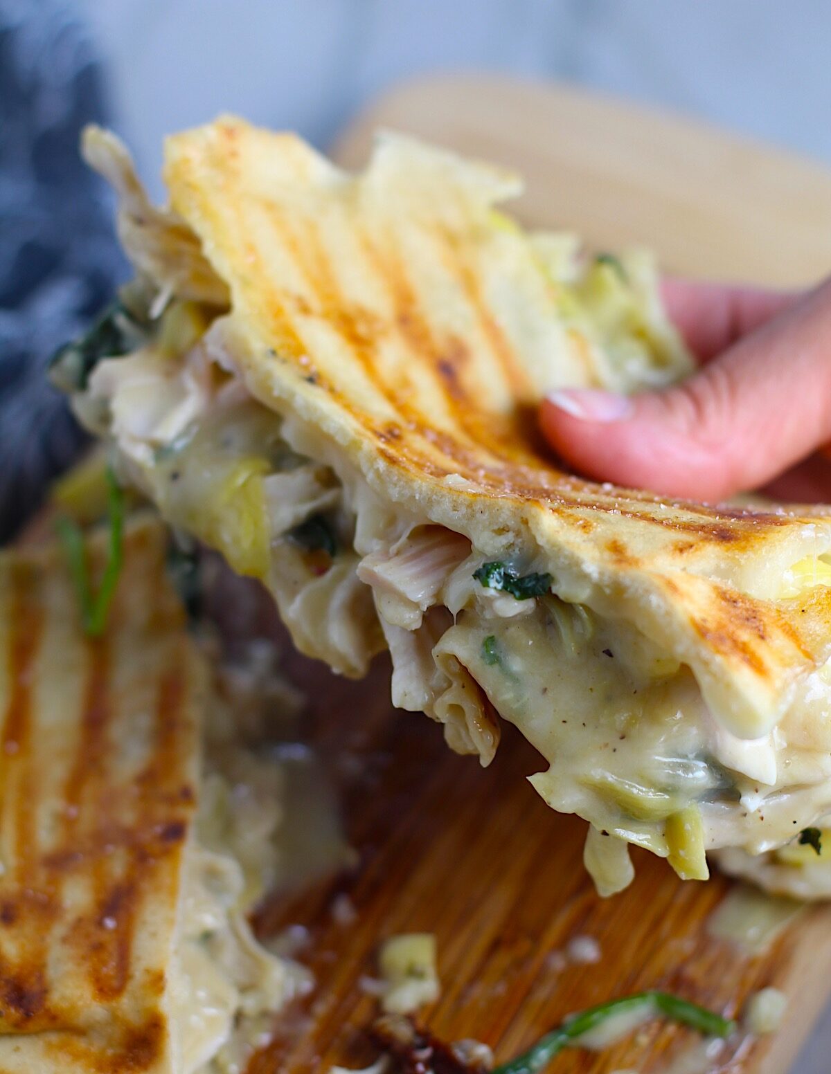 Hand holding a gooey pita sandwich with cheese and artichokes melting out. 