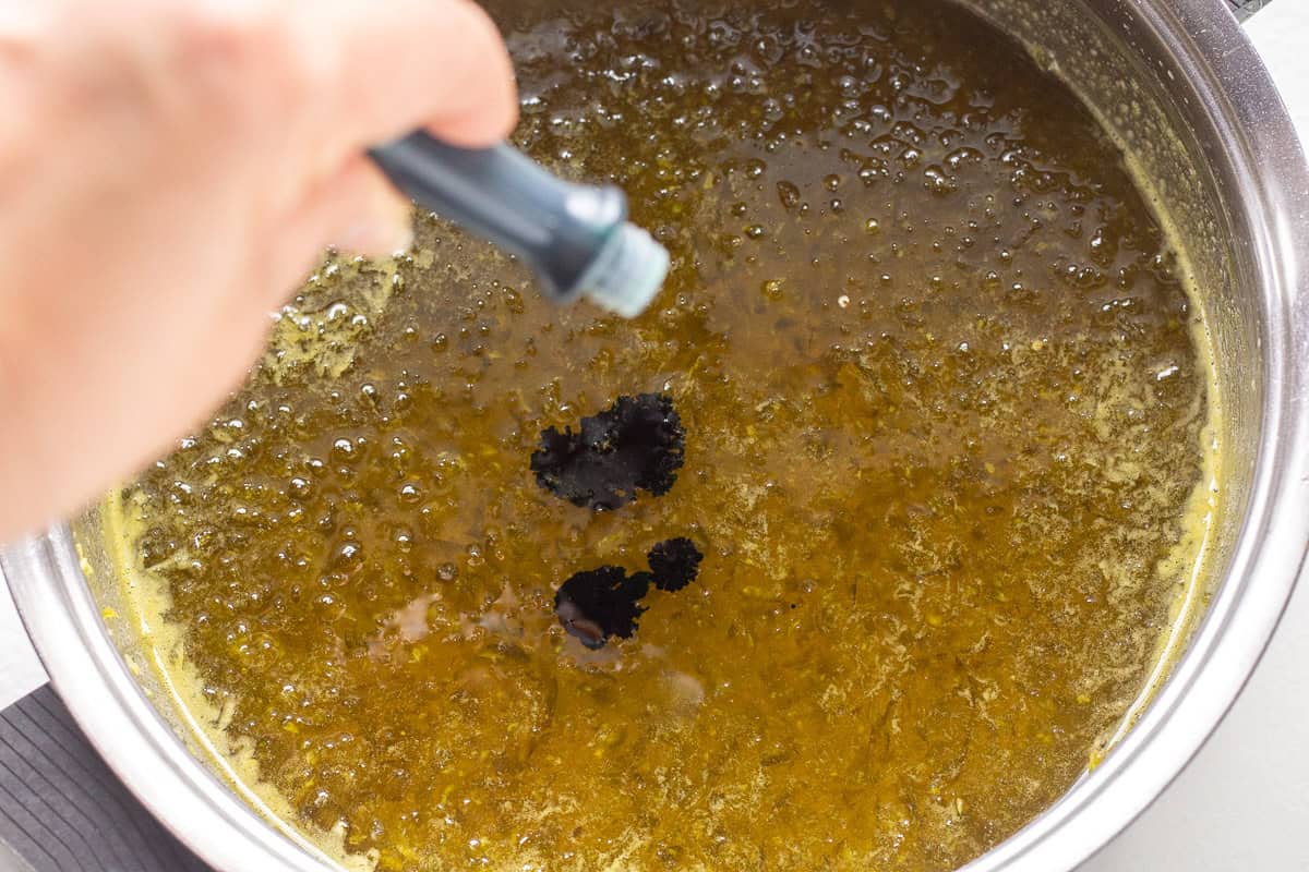 Boiling green pepper jelly in a pot with a hand adding a spice.