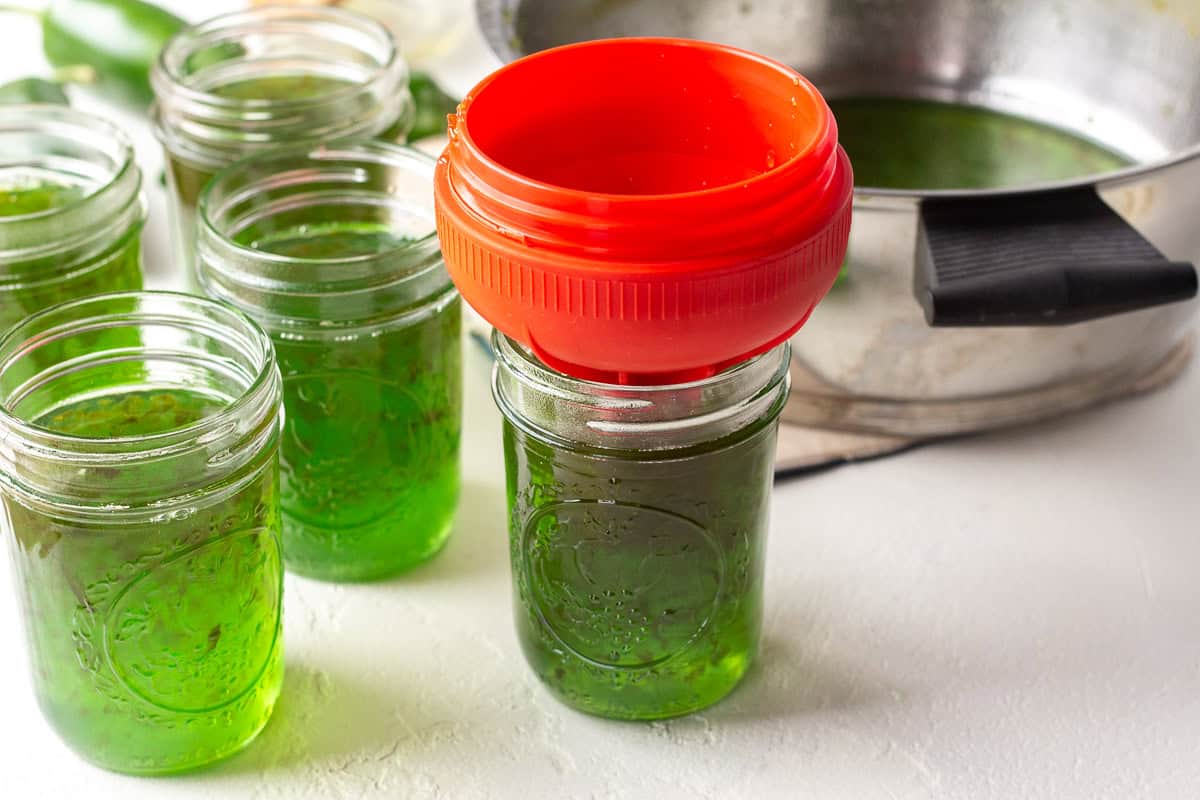 Green pepper jelly in jars with a red lid and a pot in the background.