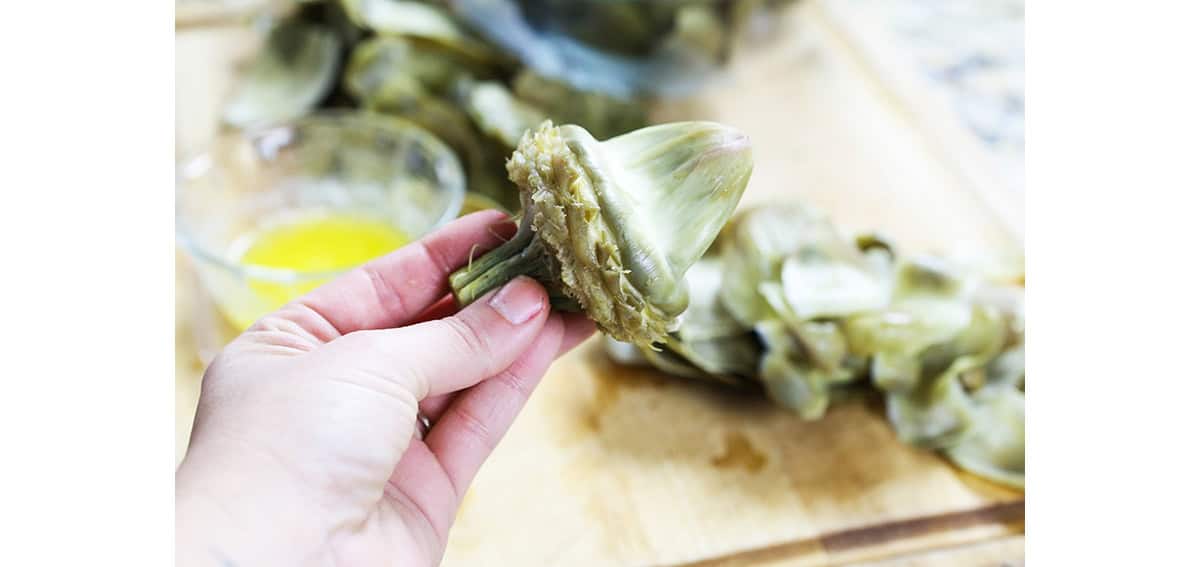 Hand holding the heart of an artichoke up above a cutting board with discarded leaves.