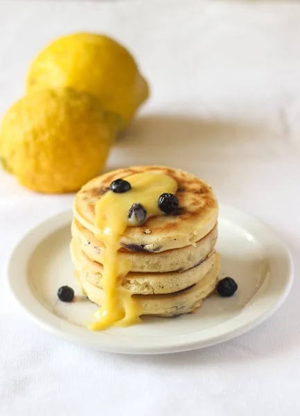 Stack of pancakes with fresh blueberries on top and lemon curd over the top and falling on the side. 