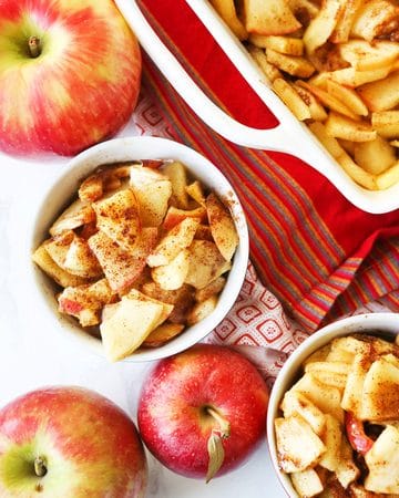 Baked apple slices with cinnamon served with fresh apples on a white surface.