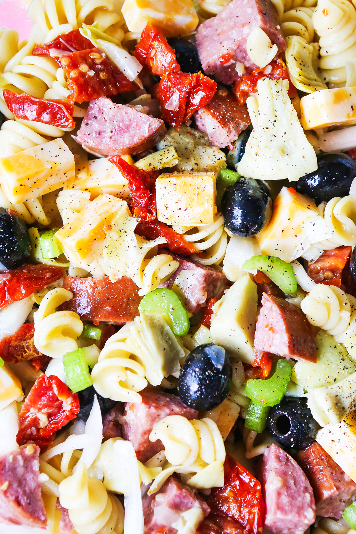 Pasta salad ingredients up close, including black olives, salami, rotini noodles, cheese and tomatoes.