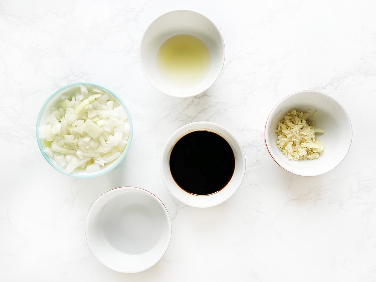Assorted bowls with chopped onions, garlic, soy sauce, and oil on a white marble surface.