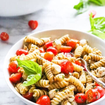 Pesto Caprese Pasta Salad with cherry tomatoes and basil in a white bowl.