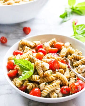 Pesto Caprese Pasta Salad with cherry tomatoes and basil in a white bowl.