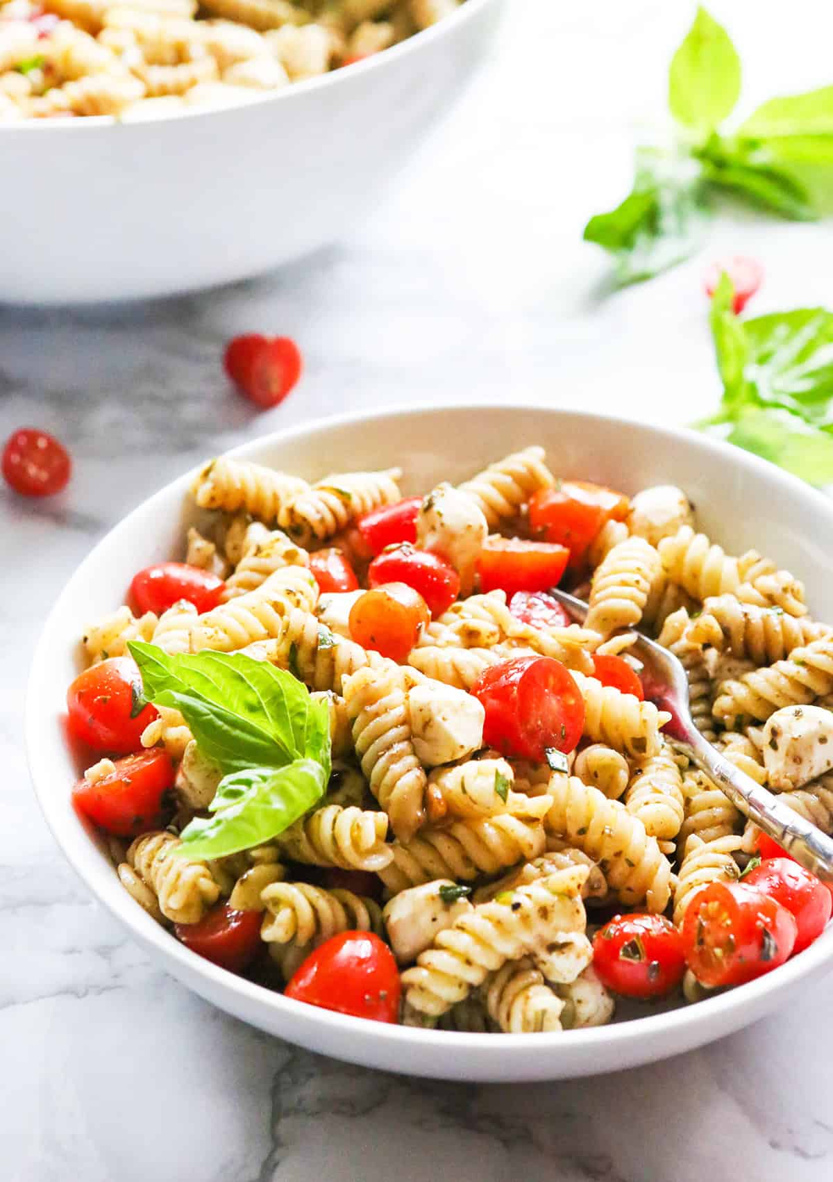 Pesto Caprese Pasta Salad with cherry tomatoes and basil in a white bowl.
