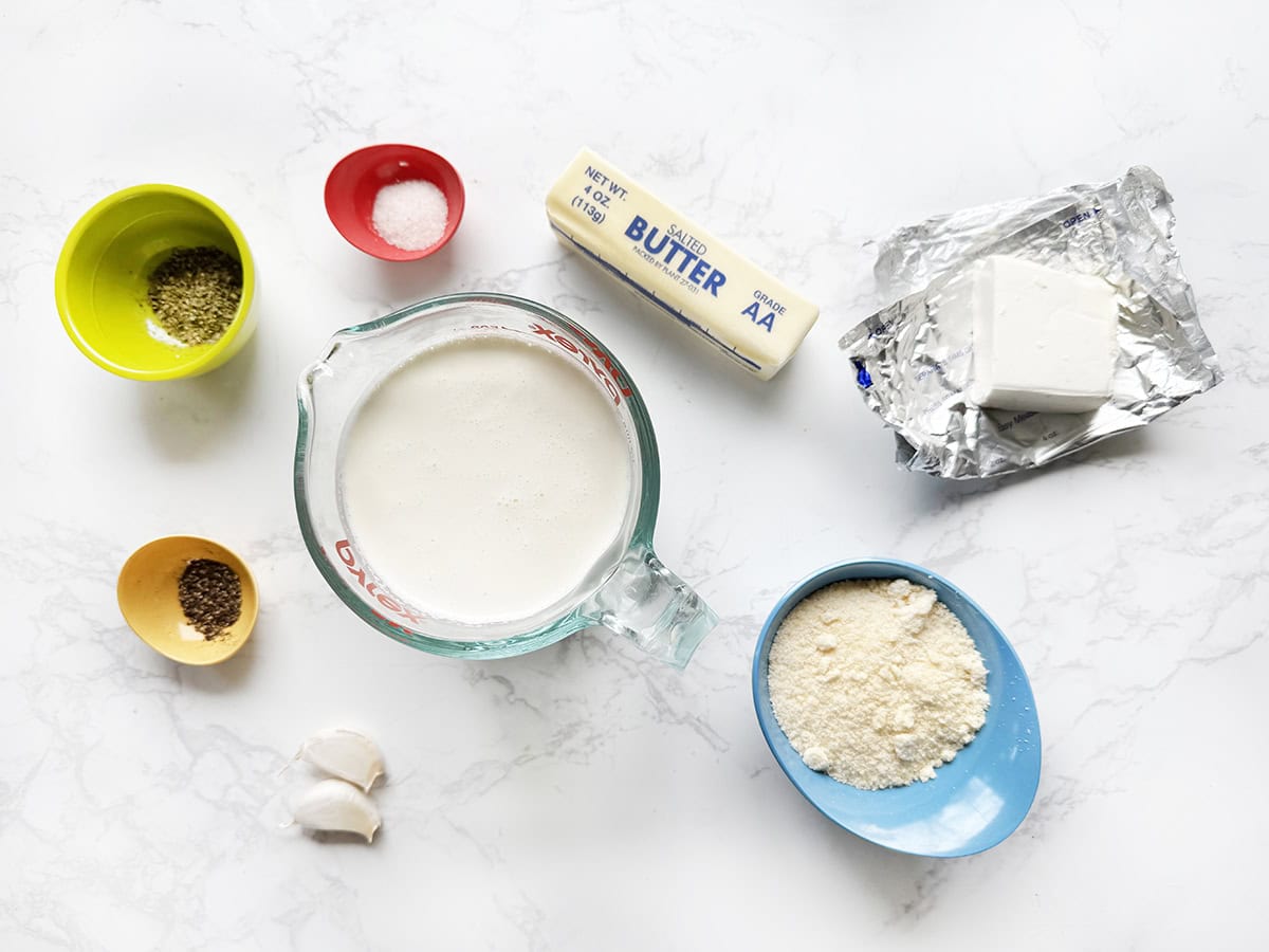 Ingredients for alfredo sauce with cream cheese in individual bowls, lined up on a white countertop.