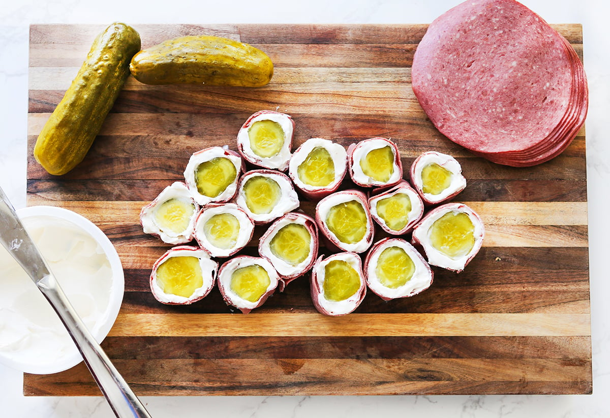Pickle rollups cut in slices and arranged on a cutting board next to pickles, corned beef and cream cheese.