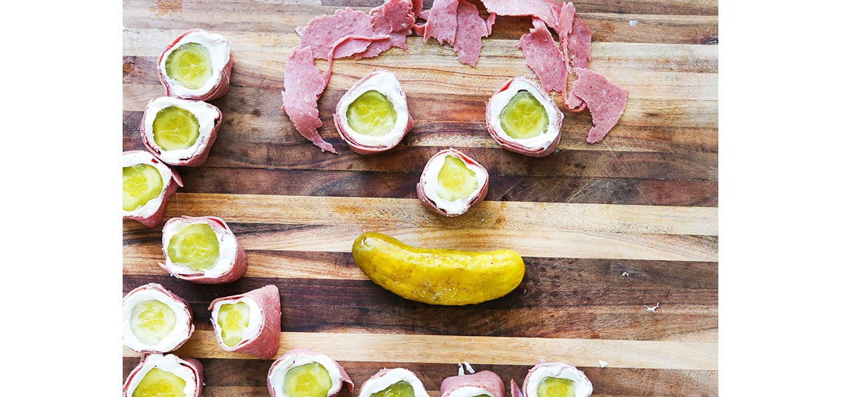 Pickle rollups and corned beef and pickles arranged in a smiley face on a cutting board.
