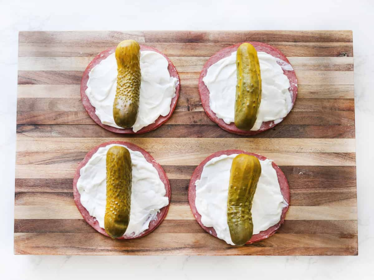 Four pickle roll ups on a wooden cutting board with cream cheese.