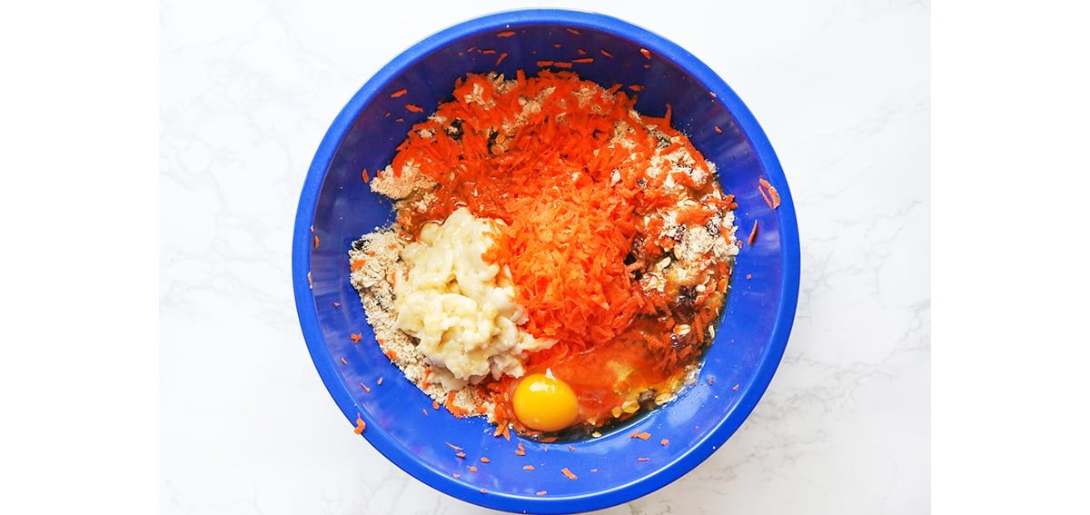Blue mixing bowl with muffin batter, shredded carrots, mashed banana and an egg.