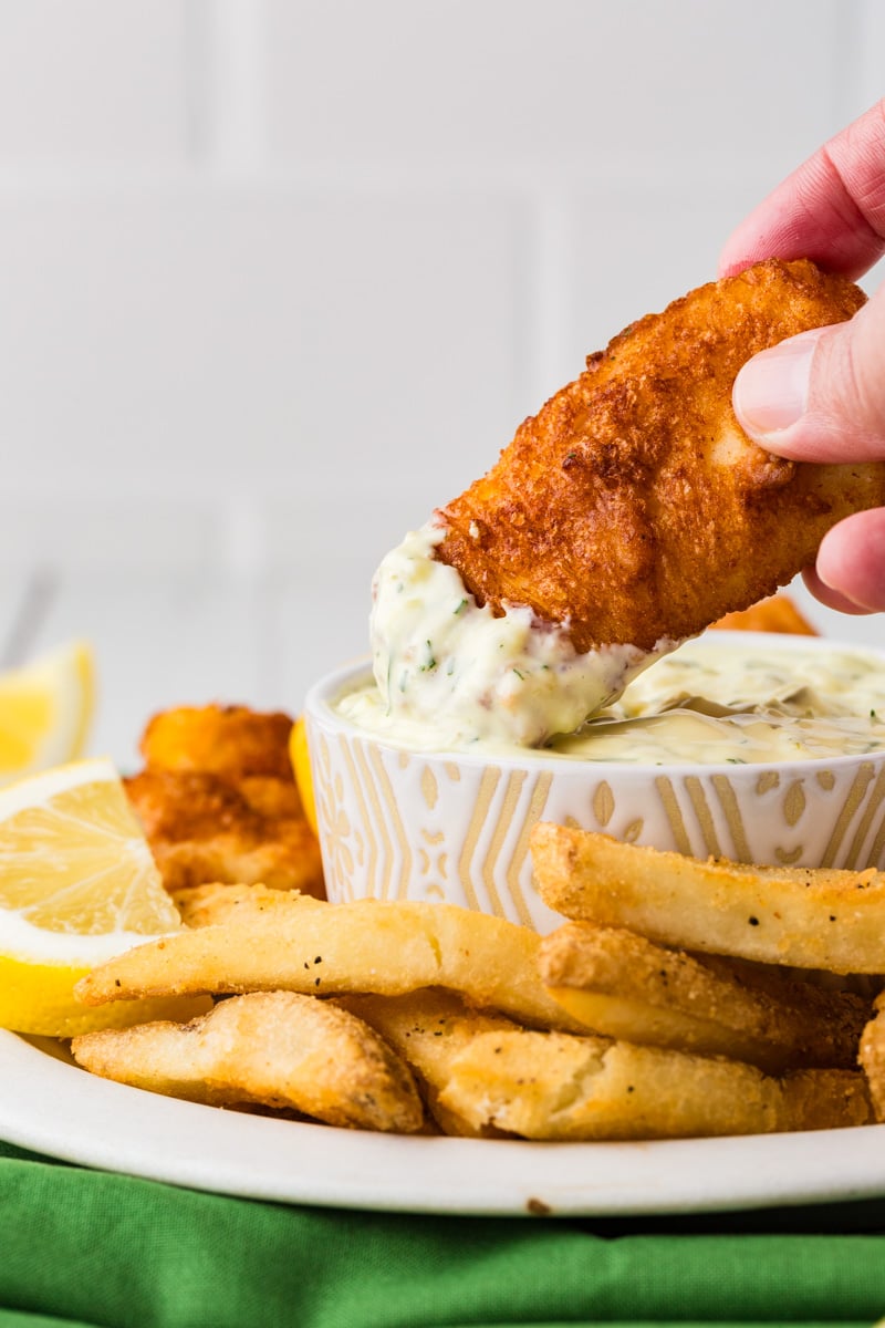 Side view of a hand dipping a fish stick into a bowl of tartar sauce.