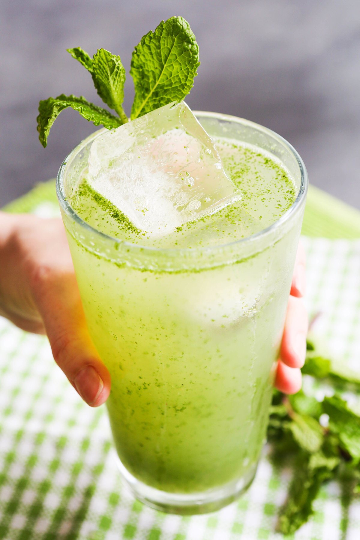 Hand holding out a green drink in a glass with a mint spring sticking out the top.