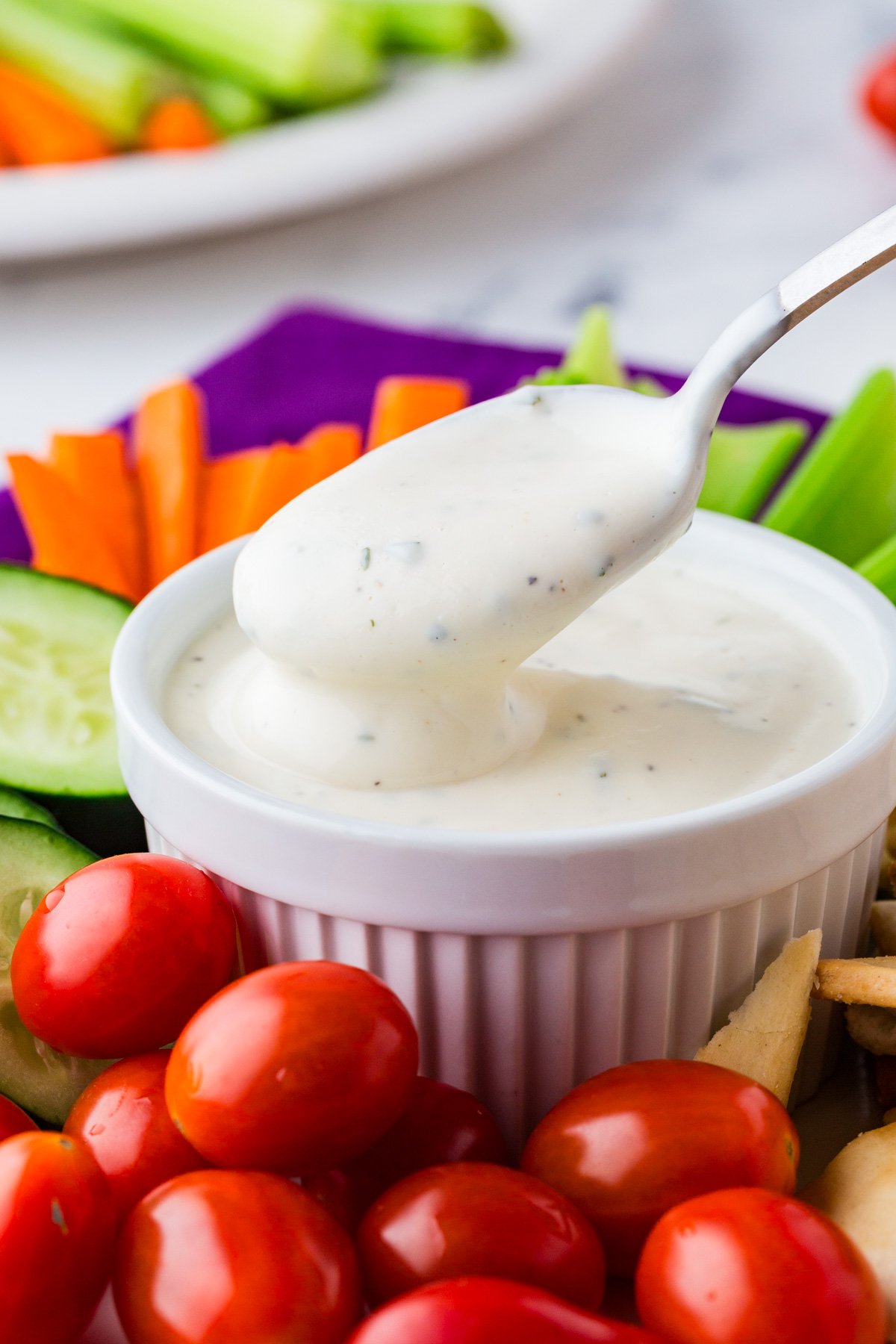 Spoon drizzling ranch dressing into a small bowl surrounded by colorful fresh vegetables.