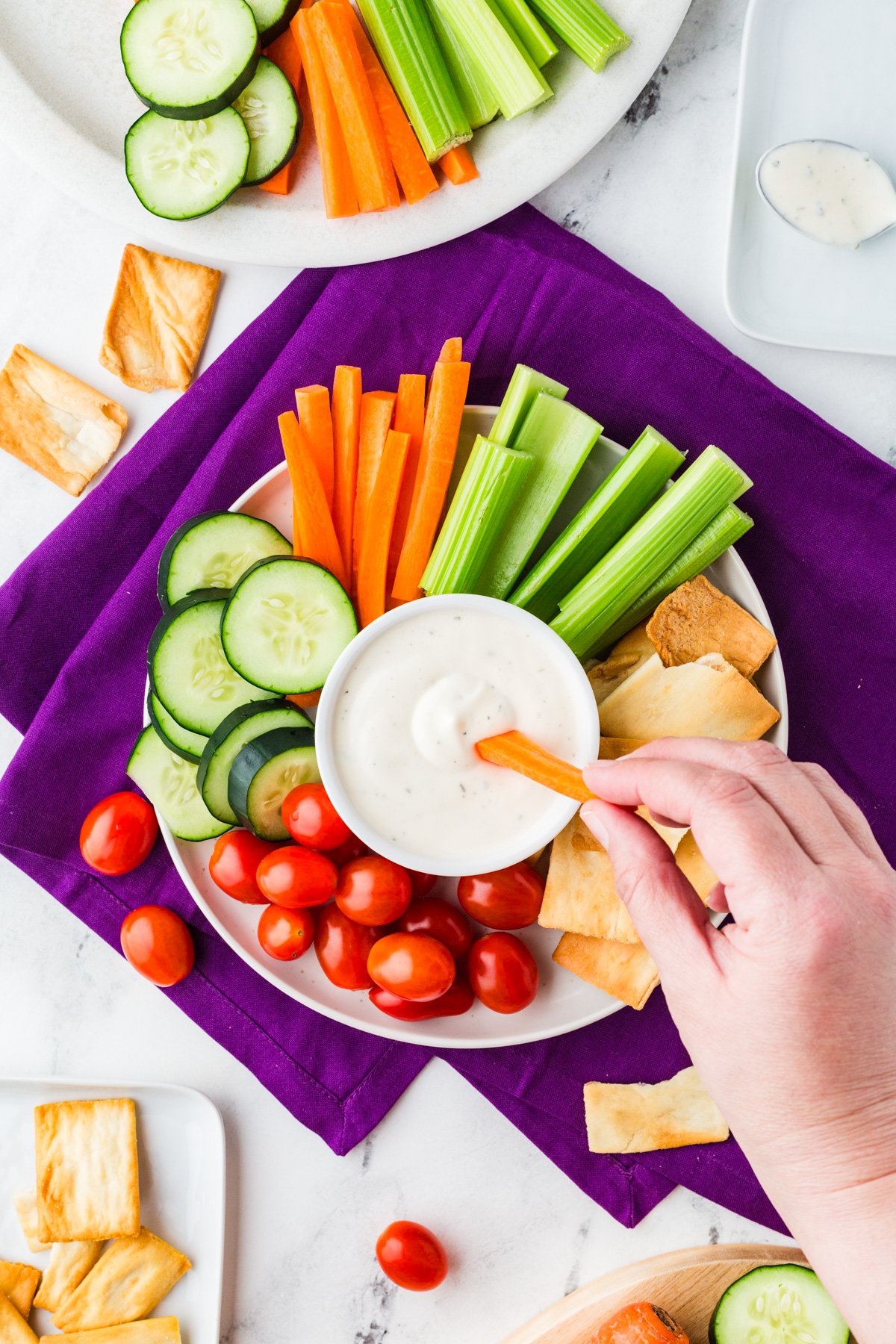 Top view of a hand dipping a carrot stick into ranch dressing, which is in the center of a veggie, cracker and dip spread.