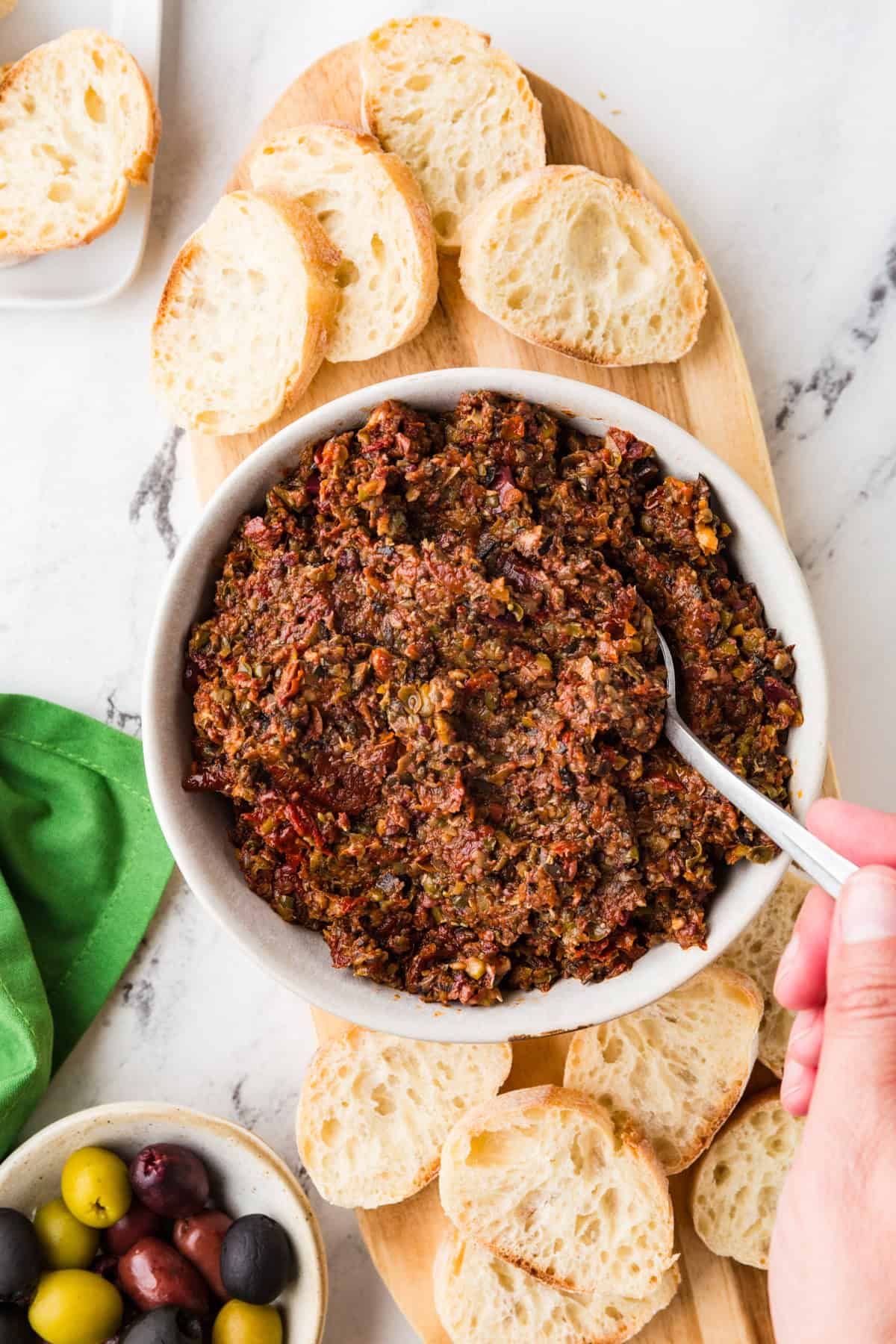 Top view of a bowl of olive tapenade with a spoon in it, surrounded by small pieces of bread.