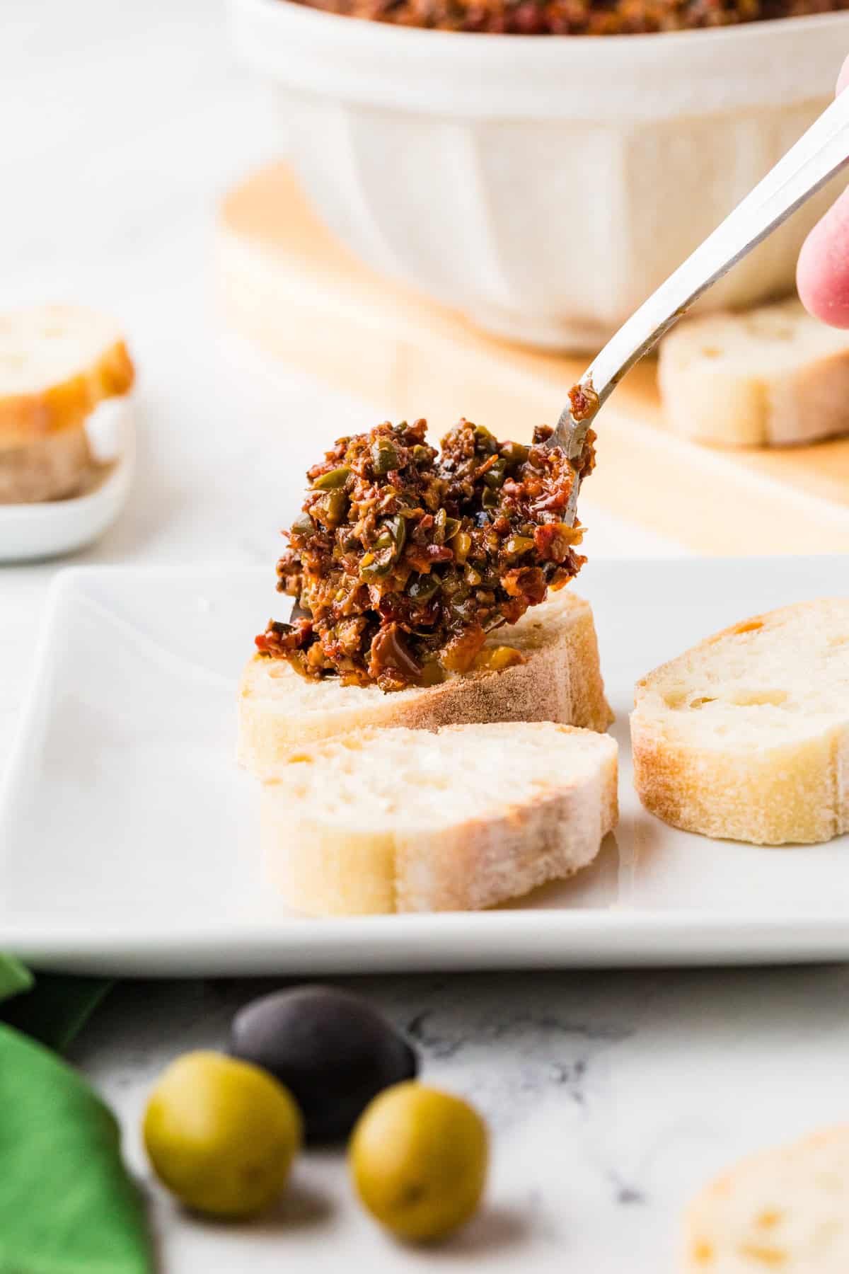 Spoon delivering some tapenade onto a piece of bread, creating what looks like bruschetta.