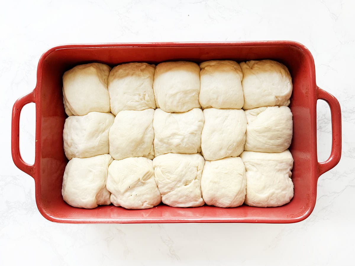 Raw dinner rolls in a red baking dish on a white surface.