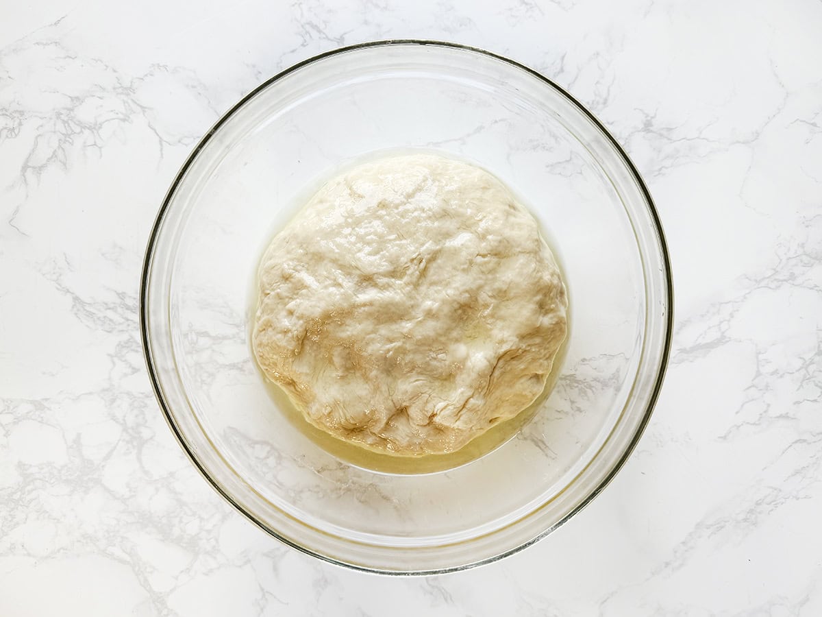 Dough rising in a glass bowl on a white marble surface.