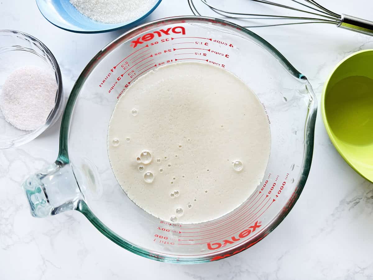Creamy liquid mixture in a glass measuring cup with baking ingredients nearby.