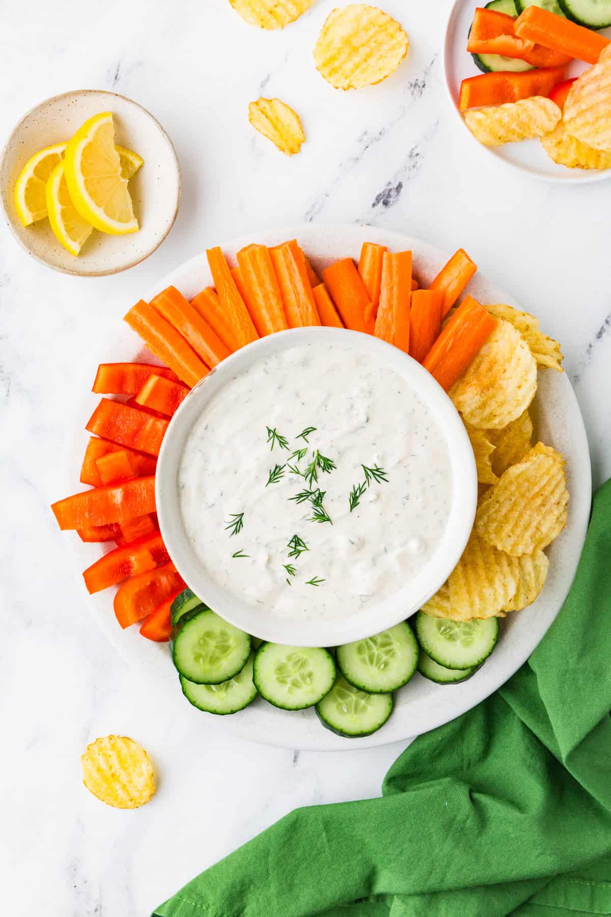 Top view of a serving platter; in the center is a creamy dill dip and it is surrounded by colorful veggies and chips.