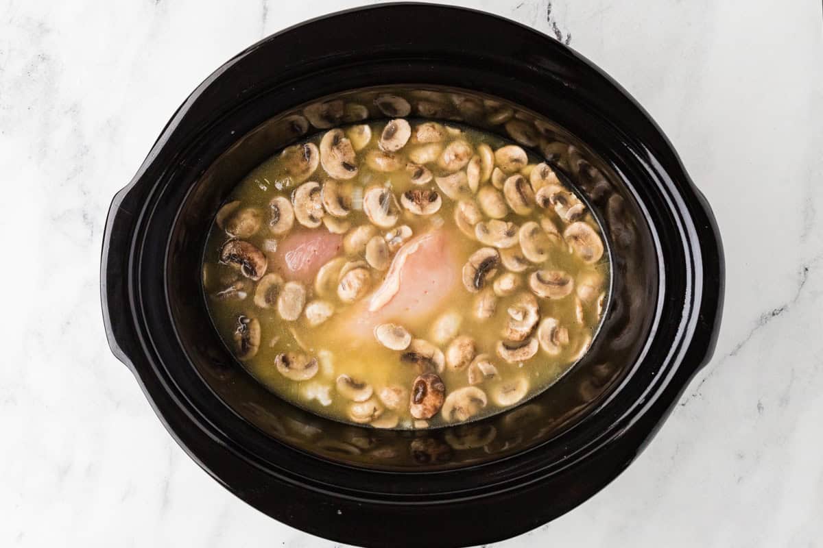 Crockpot filled with raw chicken, broth and mushrooms.