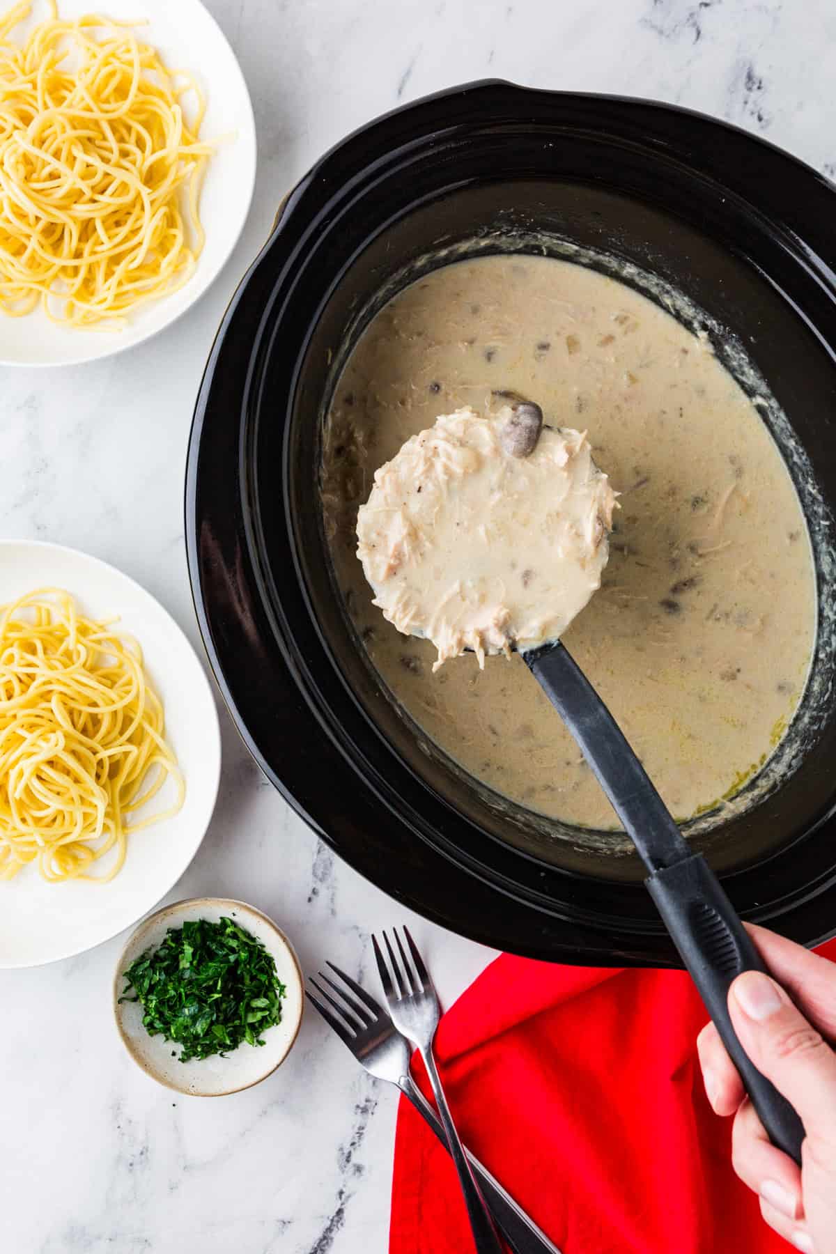 Ladle scooping chicken tetrazzini mixture out of a slow cooker, sitting next to a bowl of cooked noodles and another bowl of parsley.