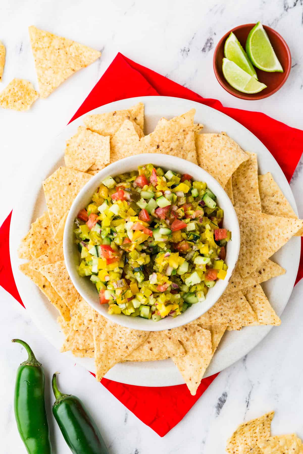 Top view of a bowl of pickle de gallo with chips around it and jalapeno peppers nearby.