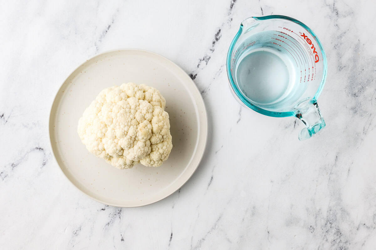 A whole head of cauliflower on a plate, next to a measuring cup holding water.