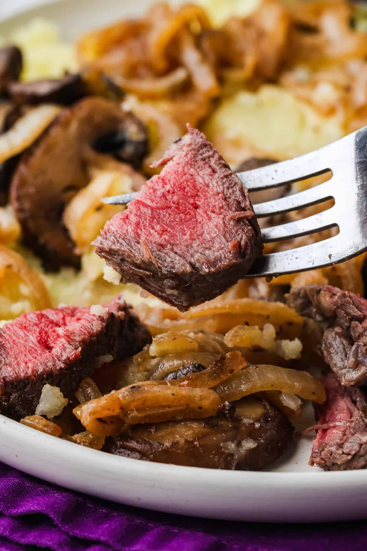 Fork holding juicy bite of steak over a plate filled with onions, mushrooms and more steak pieces.