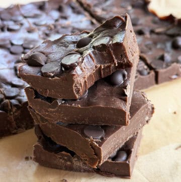 Close-up of homemade chocolate fudge topped with chocolate chips, stacked on parchment paper.
