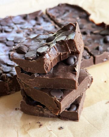 Close-up of homemade chocolate fudge topped with chocolate chips, stacked on parchment paper.