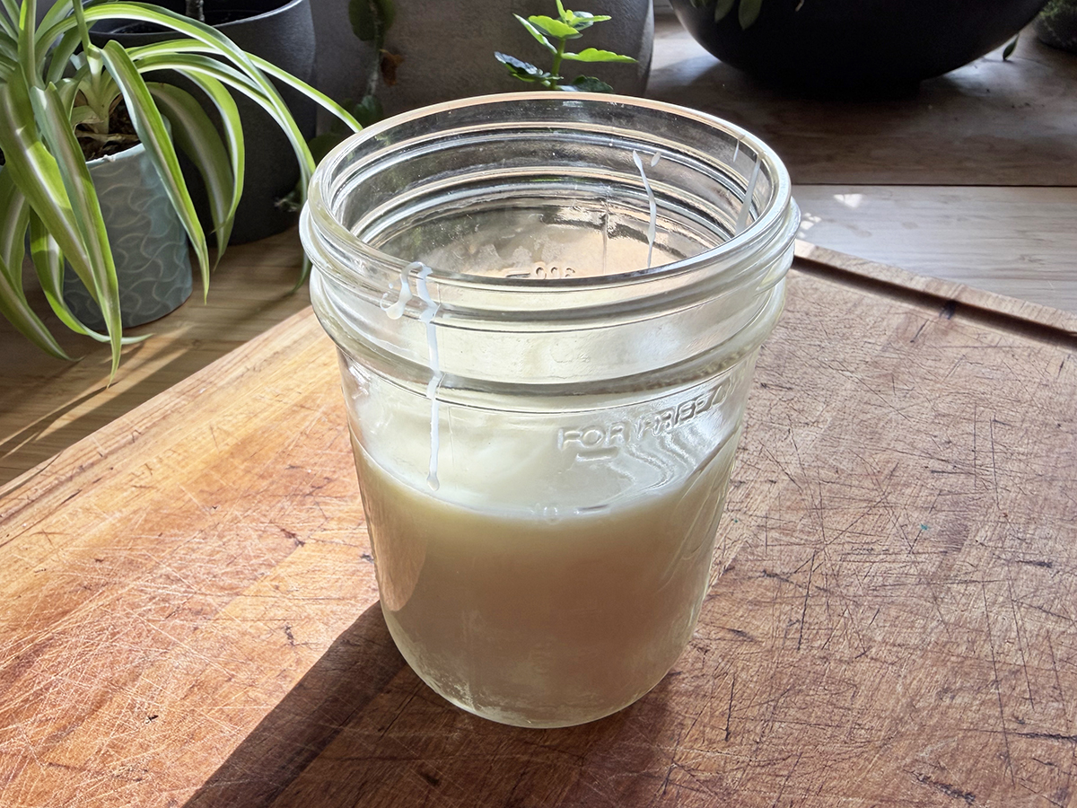 Clear homemade candle in a mason jar on wooden surface with plants in background.
