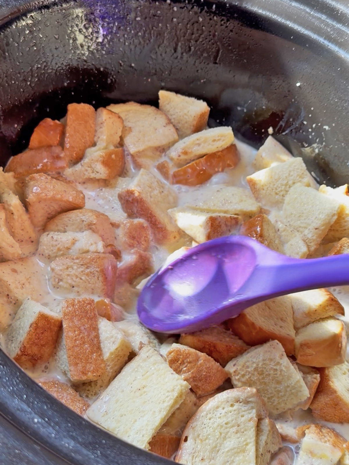 French toast cubes and apple slices cooking in a slow cooker with a purple spoon.