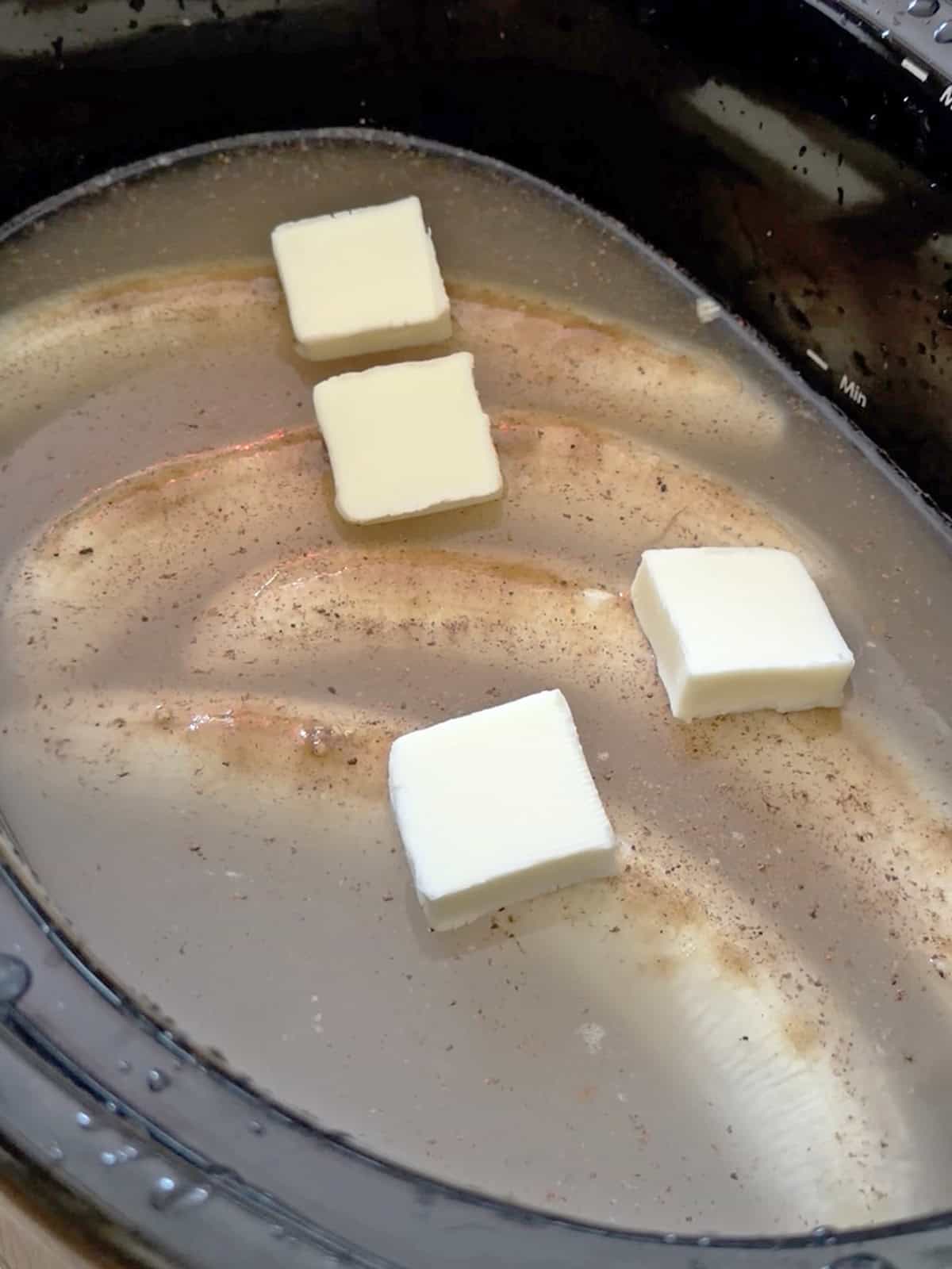 Four butter cubes melting in a slow cooker with liquid and cinnamon.