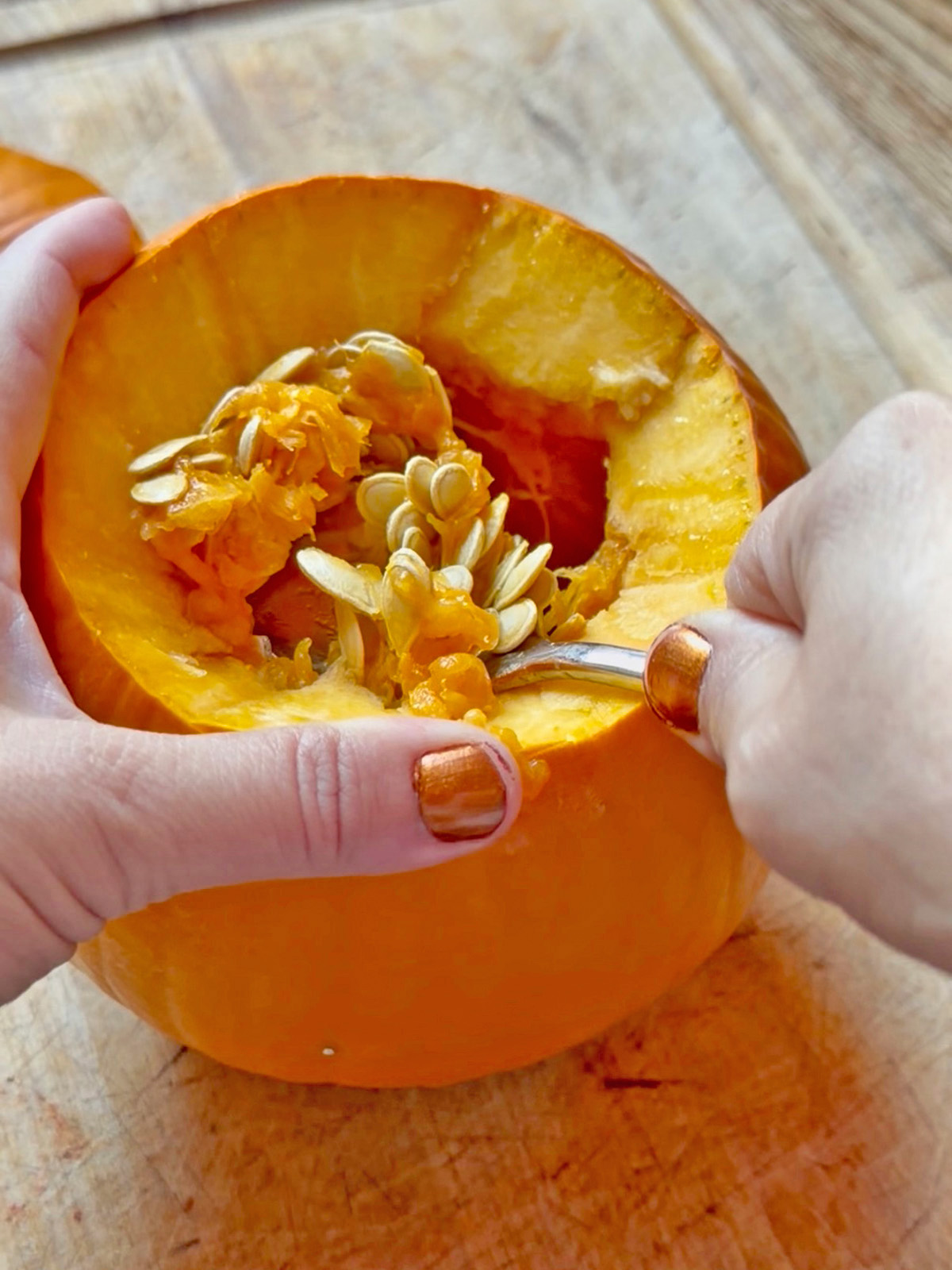 Scooping pumpkin seeds from a hollowed pumpkin with a spoon on a wooden surface.