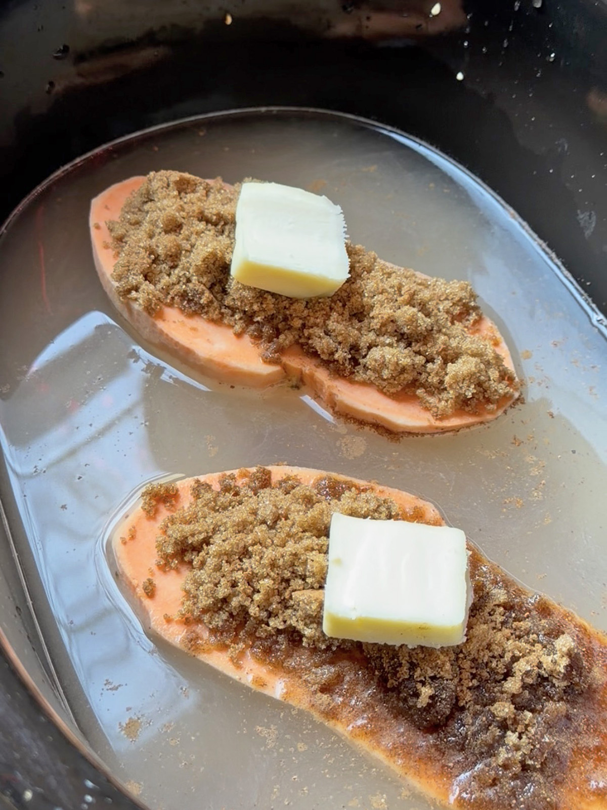 Two slices of sweet potato topped with brown sugar crumble and a pat of butter in a slow cooker.