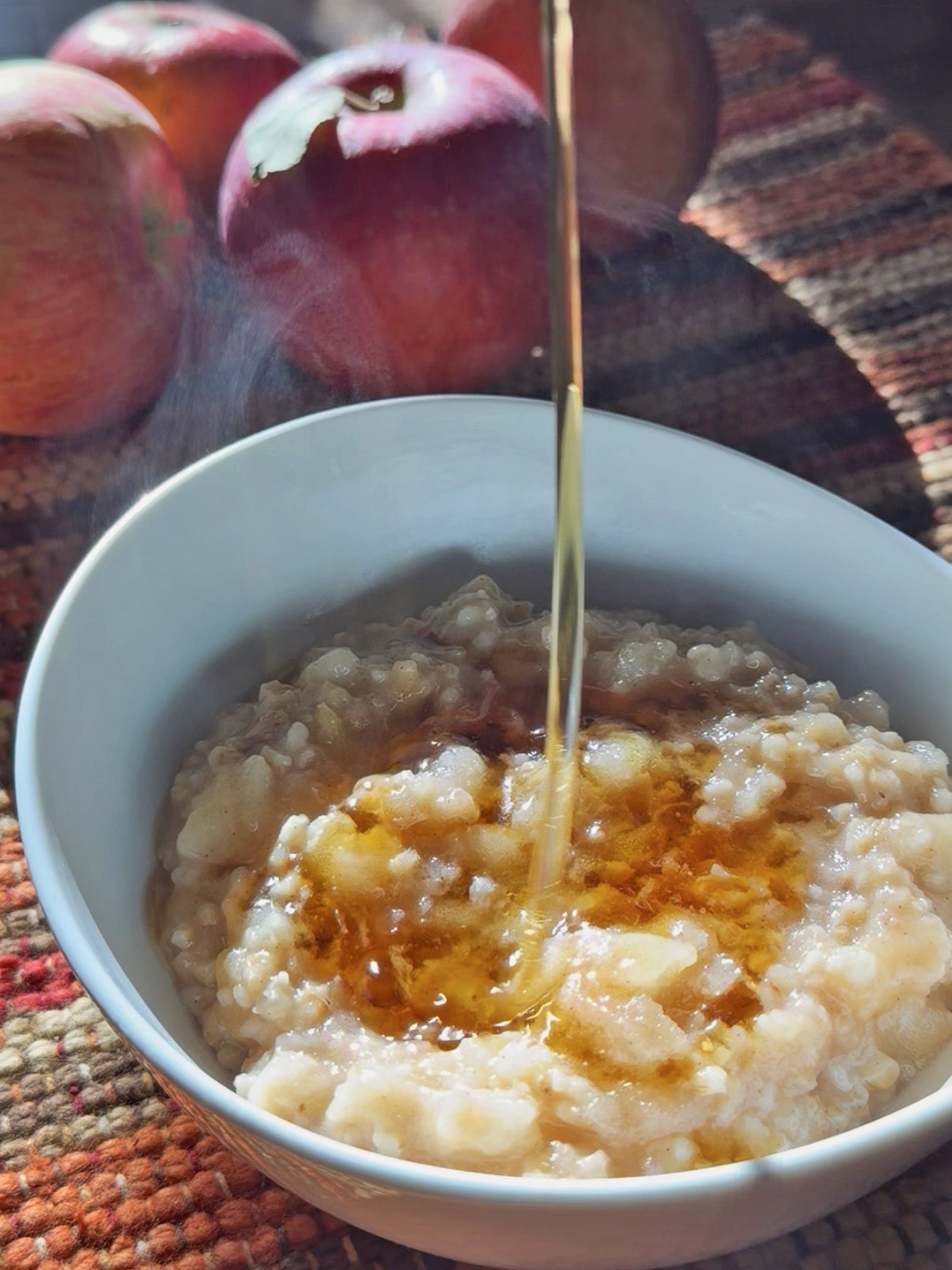 Warm apple oatmeal topped with syrup in a white bowl, with apples in the background.